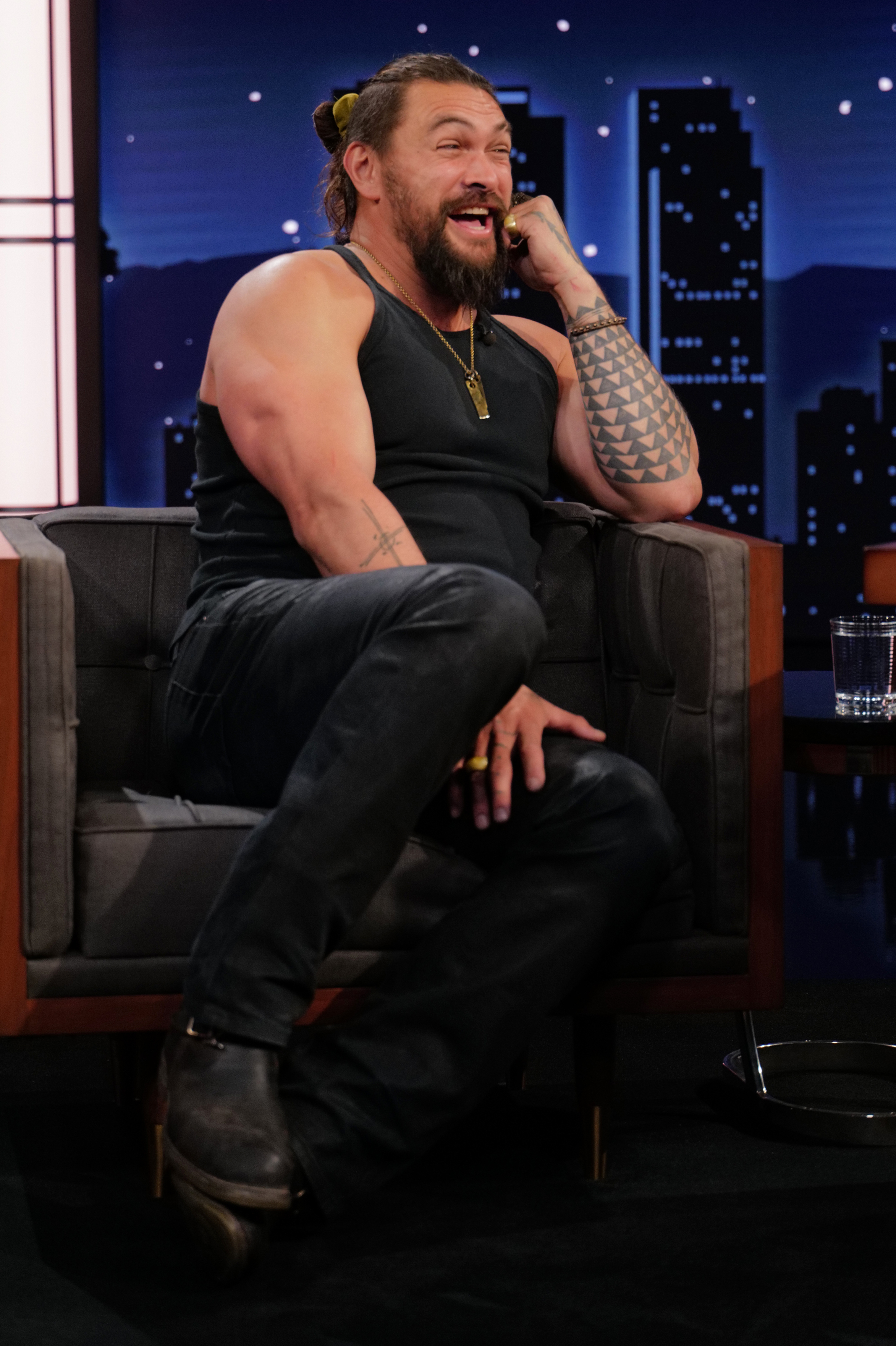 Jason Momoa seated in a talk show setting, wearing a tank top and jeans, laughing with a hand to his ear