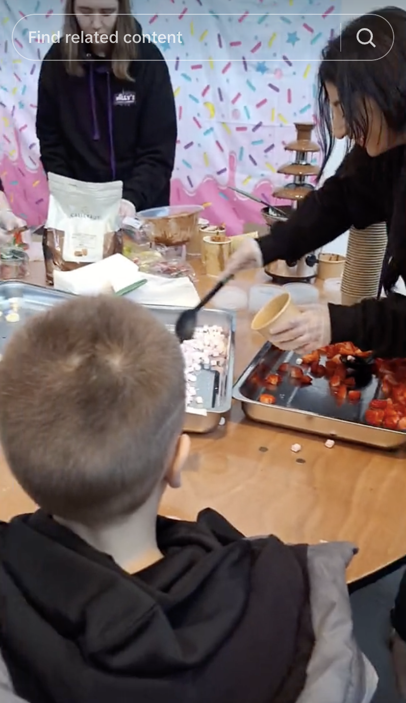 Child watches as an adult prepares food with strawberries and chocolate at a table