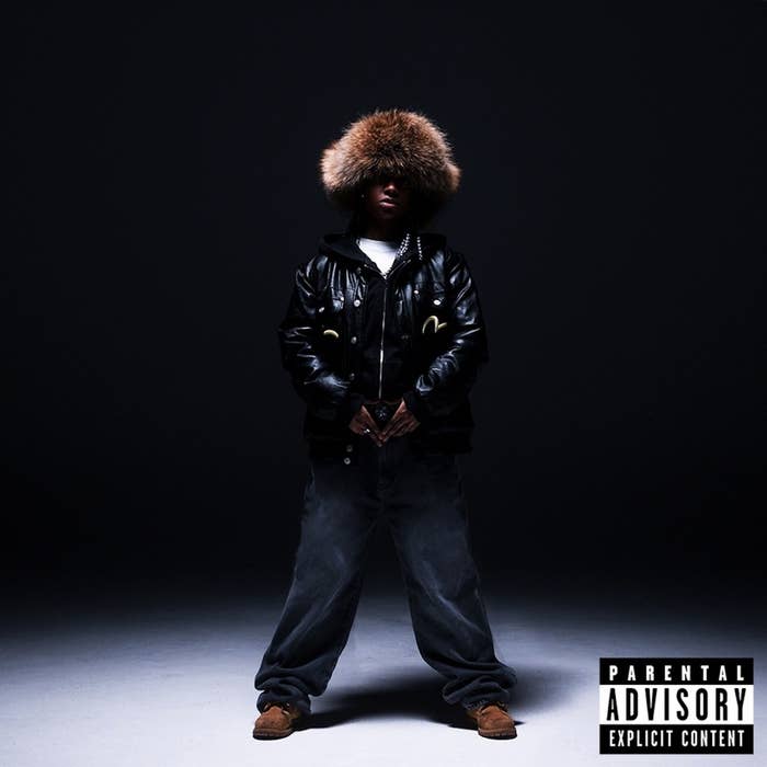 Music artist standing with hands in pockets, wearing a fur-trimmed hooded jacket, and a Parental Advisory label visible