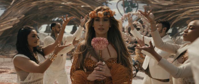Jennifer in an ornate costume with a floral headpiece surrounded by dancers in a choreographed pose in a scene from the musical movie