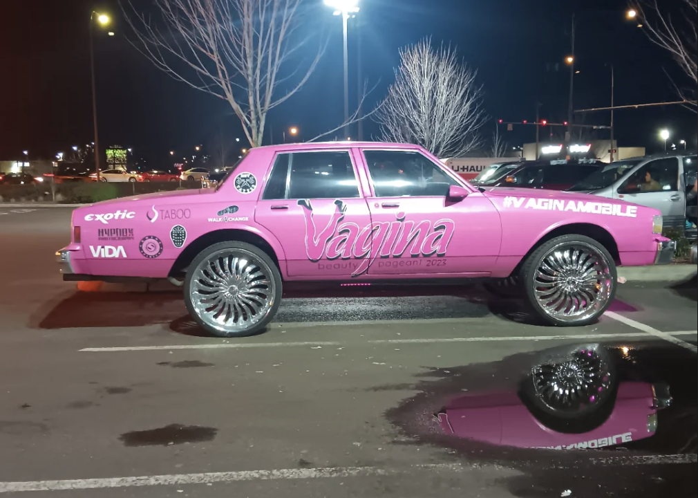 A pink car with vagina prominently written on its side is parked in a lot at night
