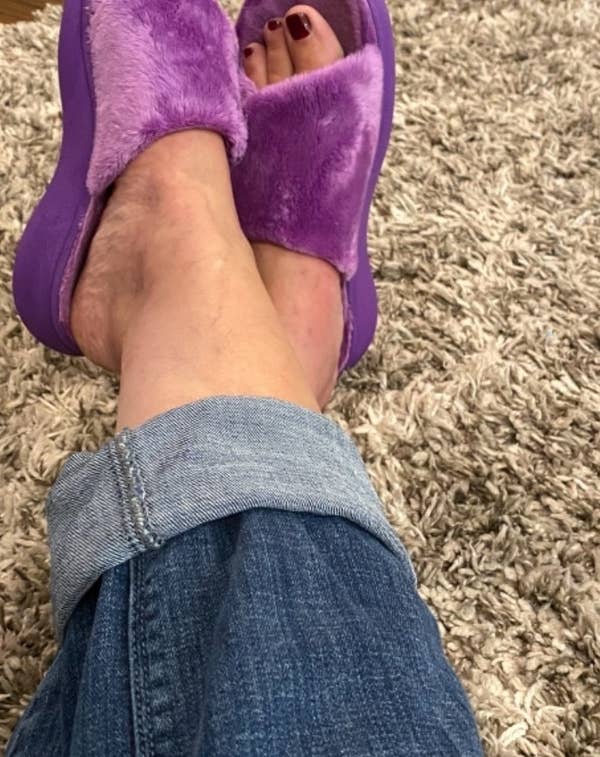 reviewer photo of feet in plush purple slippers with denim jeans on a shaggy rug