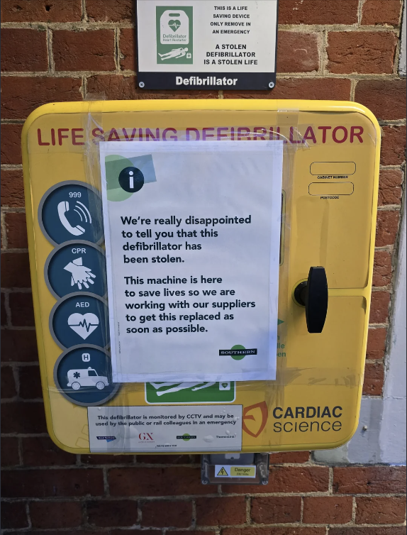 Defibrillator station with a notice about the machine being stolen and expressing disappointment. Signs include emergency instructions