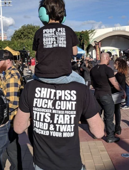 Child on parent&#x27;s shoulders; both wear profanity-laden shirts at an event. Shirt text reflects bond and humor