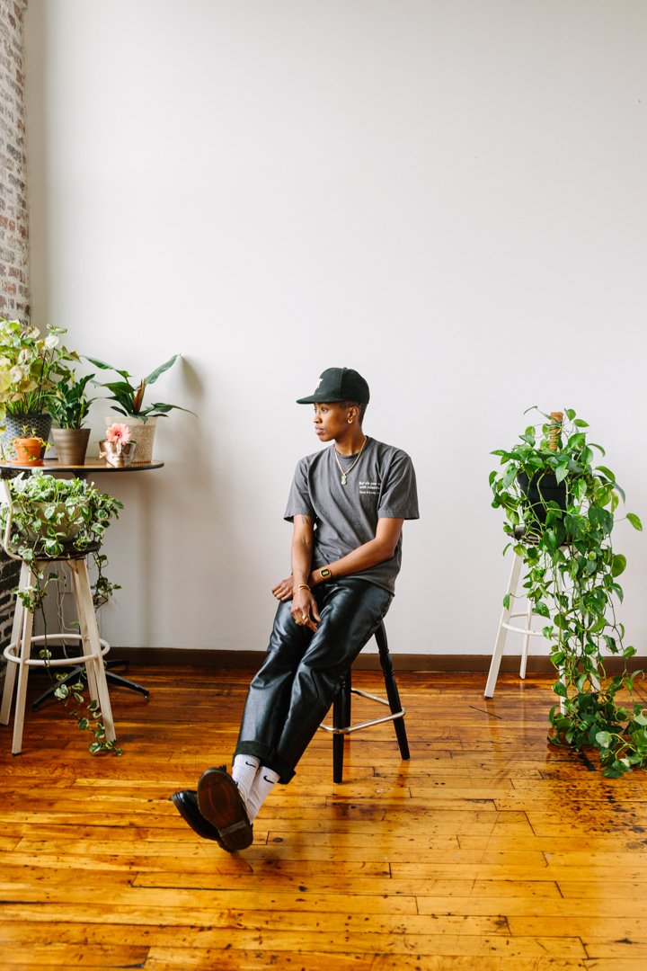 Mal Wright sitting on a chair beside houseplants, wearing a cap, T-shirt, and loose pants