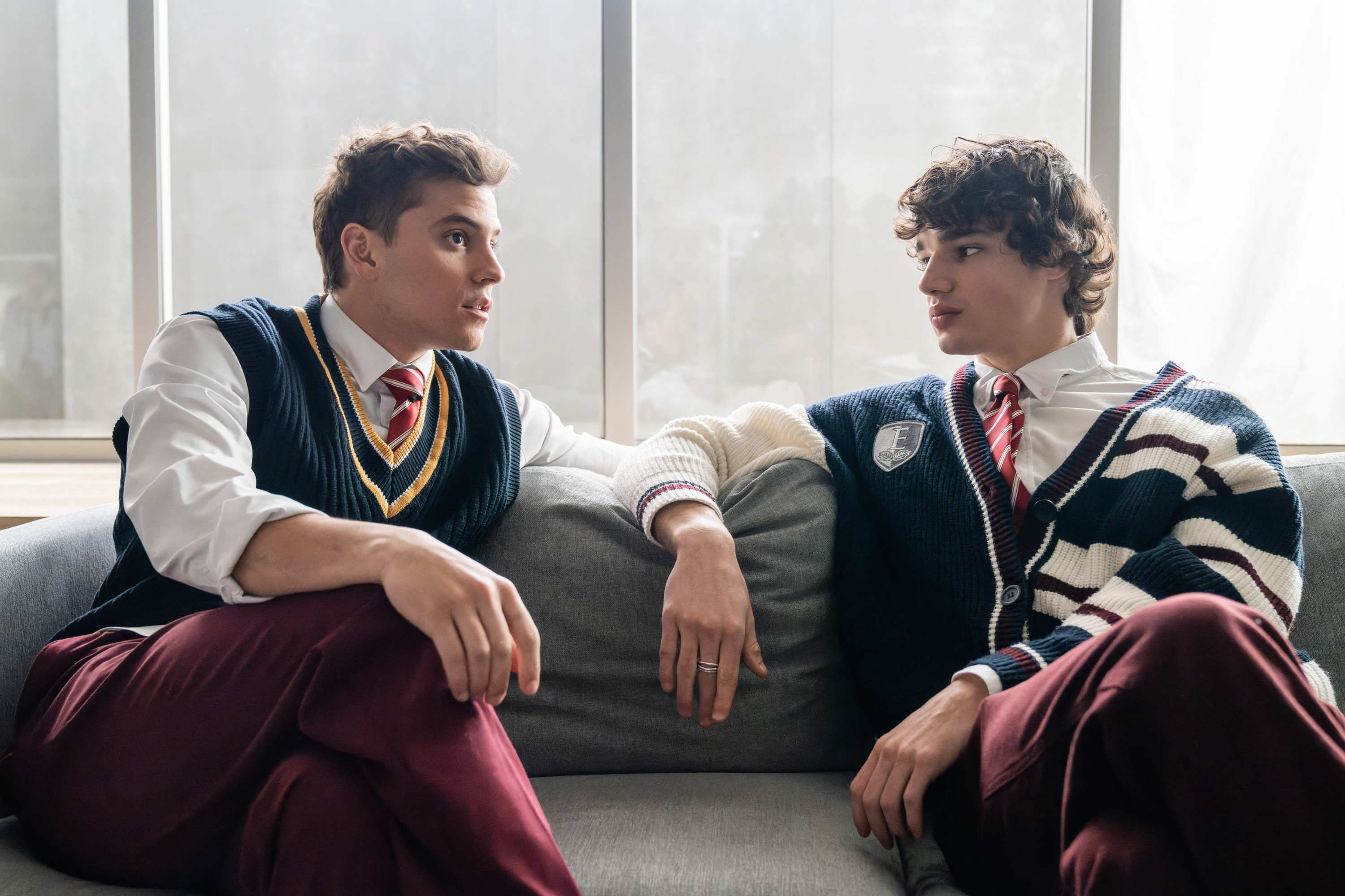 Two actors in preppy outfits sitting on a couch, engaging in a conversation for a TV show scene