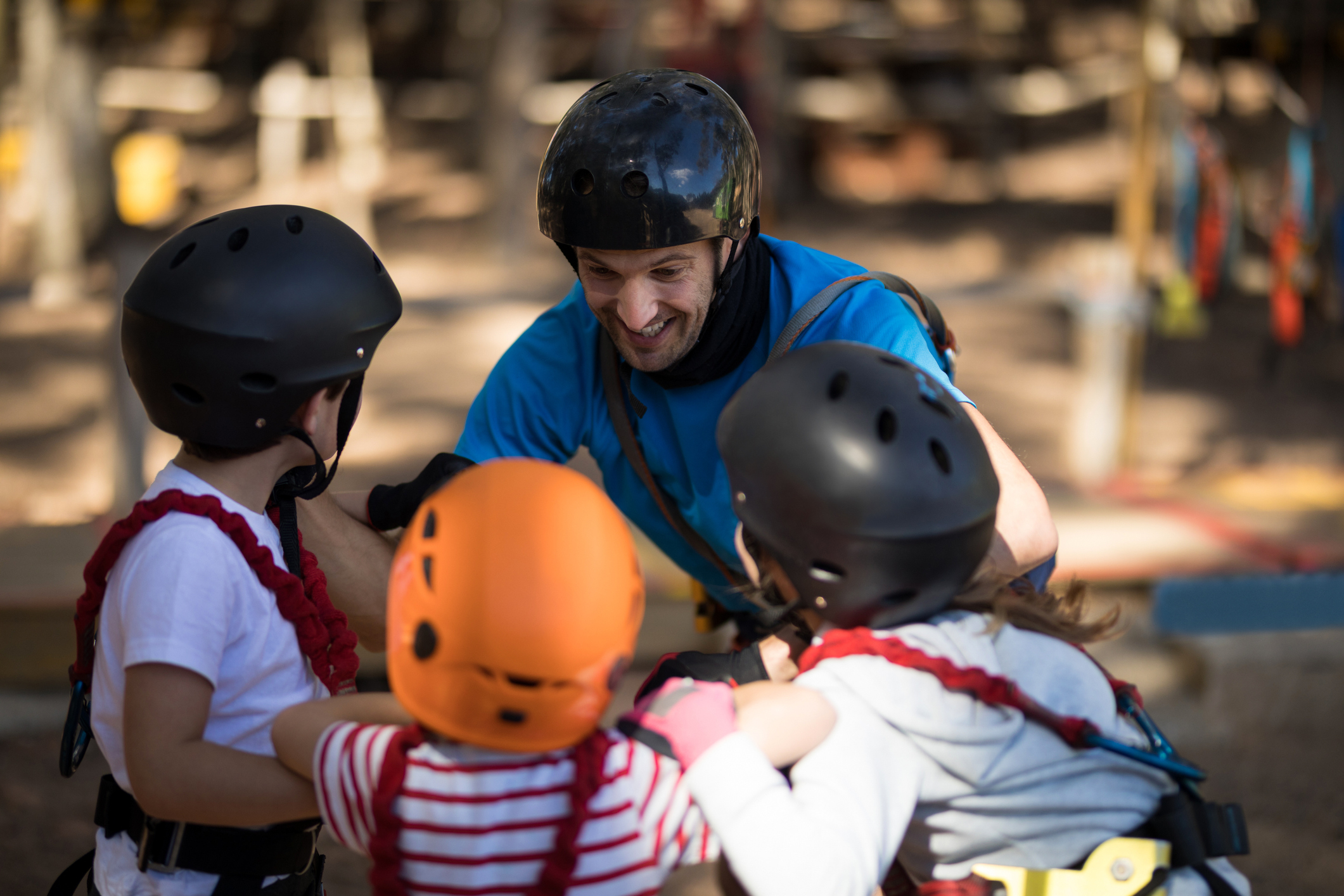 Instructor in a blue shirt secures harnesses on two children wearing helmets before a ropes course