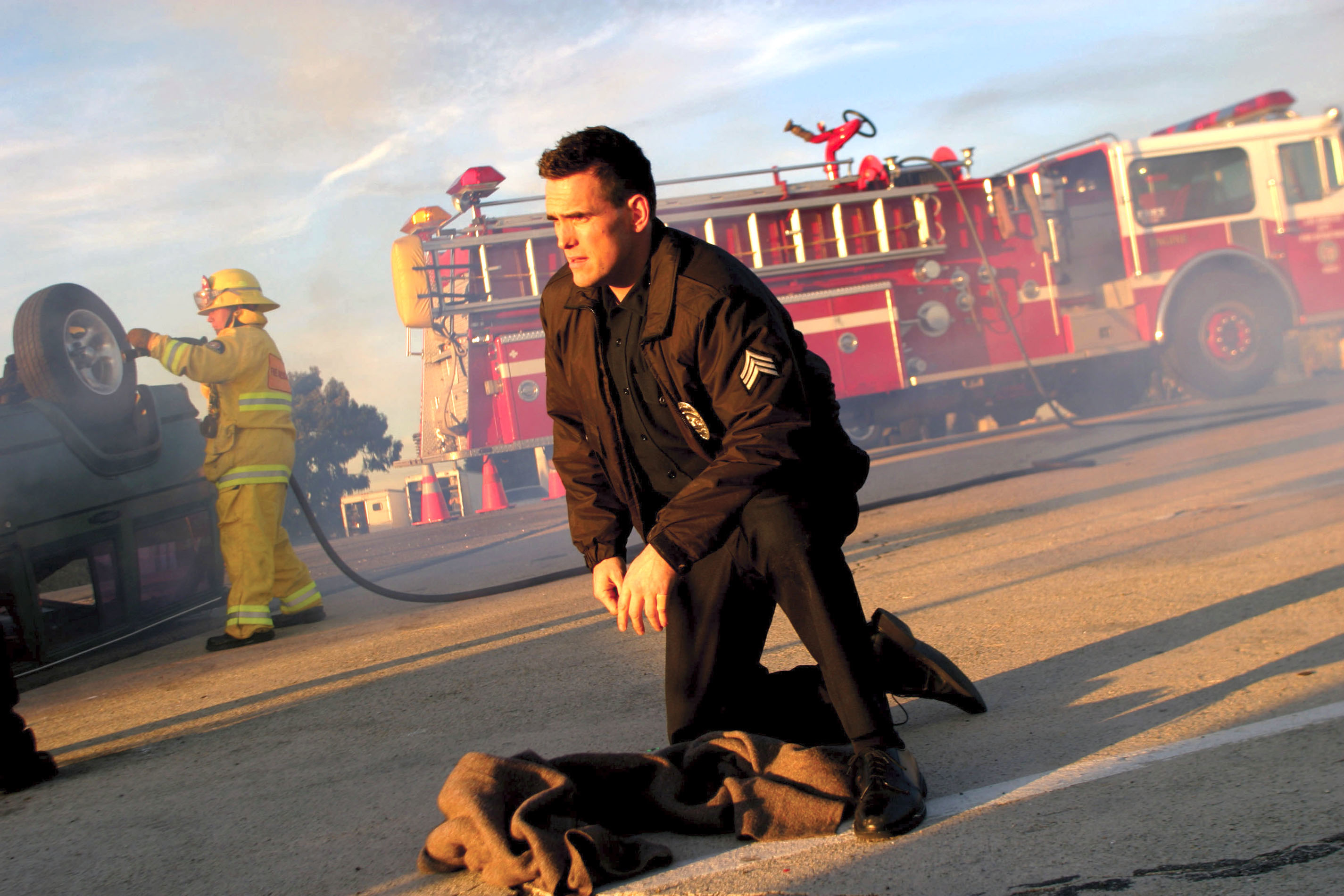 Firefighters in action, Matt Dillon as Officer John Ryan, kneeling in foreground with equipment, a fire truck, and a colleague in background