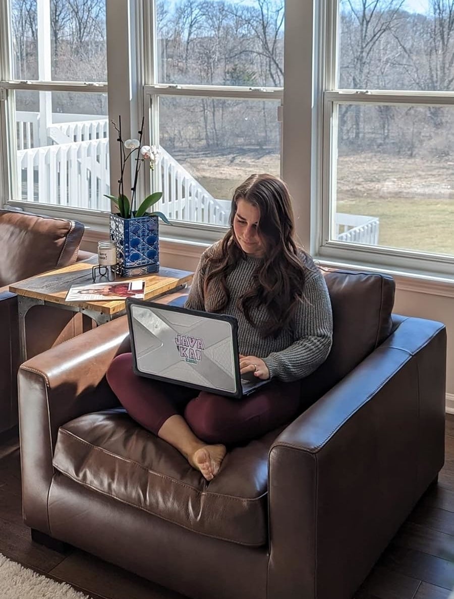 Person on a brown chair working on a laptop, with a book and vase on the table, indoors with a view of leafless trees outside