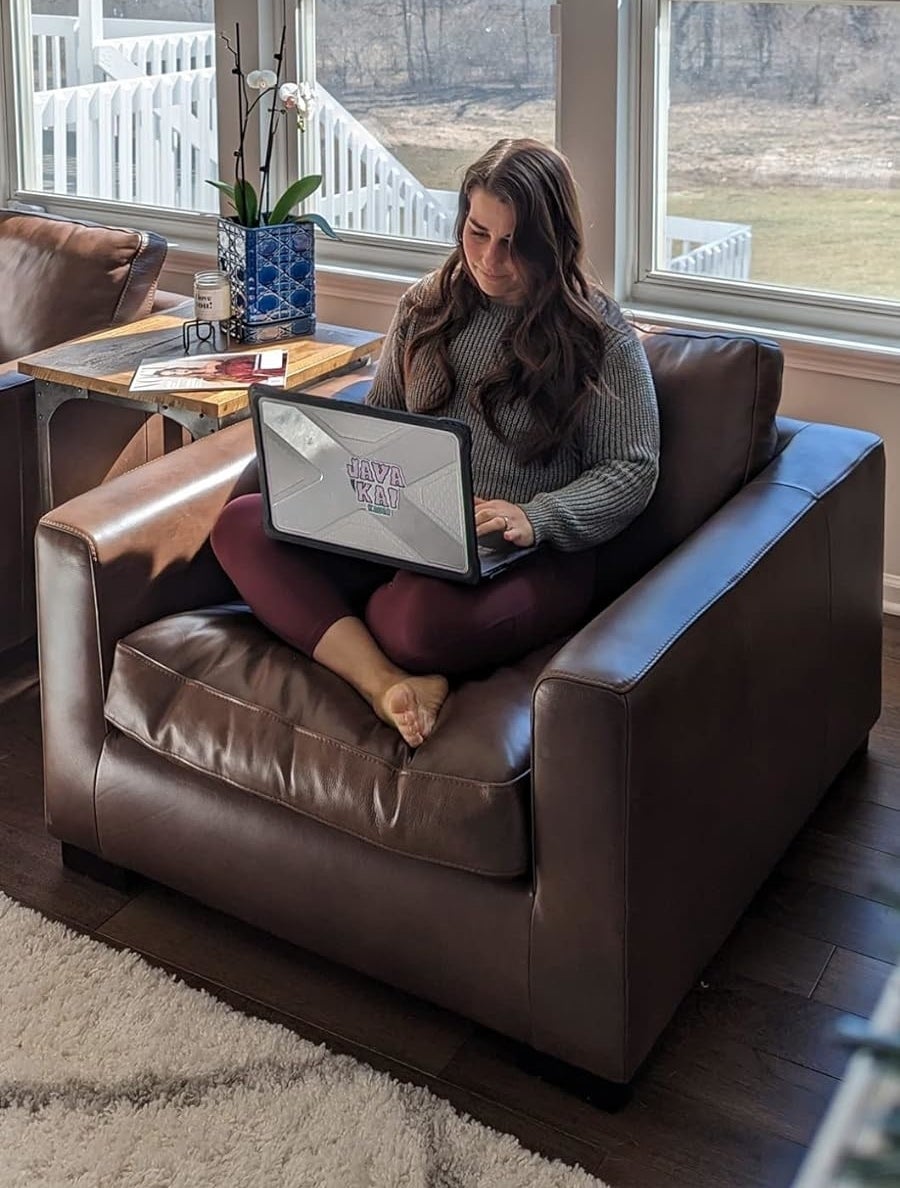 Person on a brown chair working on a laptop, with a book and vase on the table, indoors with a view of leafless trees outside