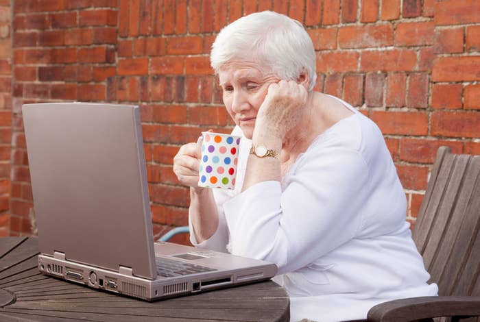 Elderly woman using a laptop outdoors, holding a mug. She appears thoughtful or concerned