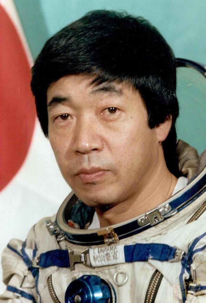 Toyohiro in a spacesuit with mission insignia, against a flag background