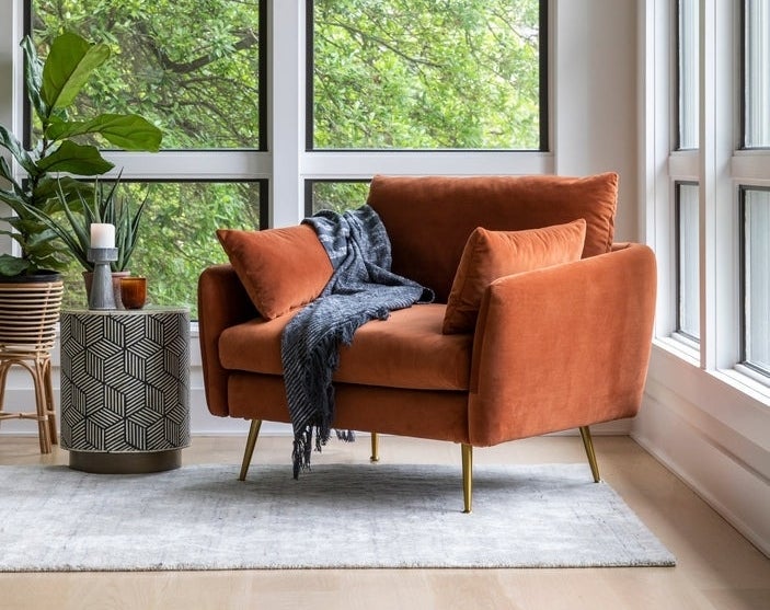 Stylish room with a trendy burnt orange chair, geometric side table, and cozy grey throw, alongside large windows with a leafy view
