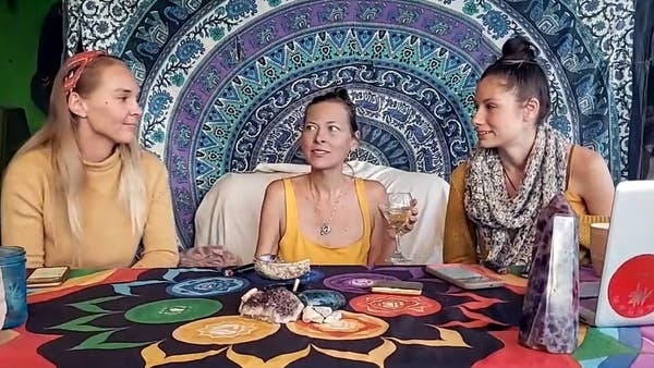 Three individuals at a table with a tapestry backdrop, crystals, and tarot cards. They seem engaged in a spiritual or mystical session