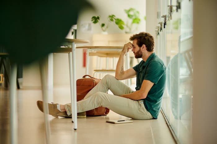Man sitting on floor by window, resting head on hand with bag beside him, appears stressed
