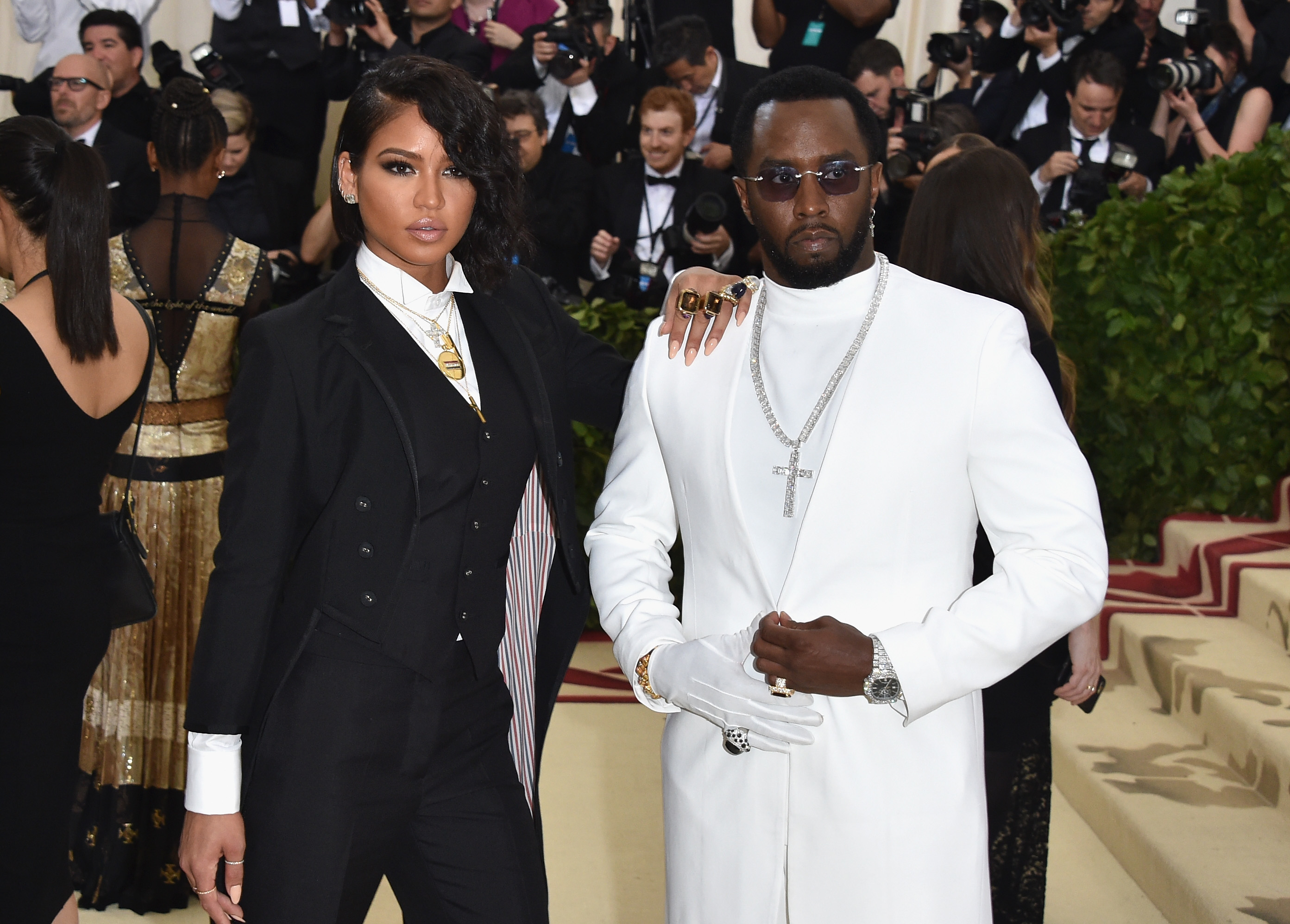 Two stylish individuals posing at an event, one in a white suit and the other in a dark ensemble with unique accessories