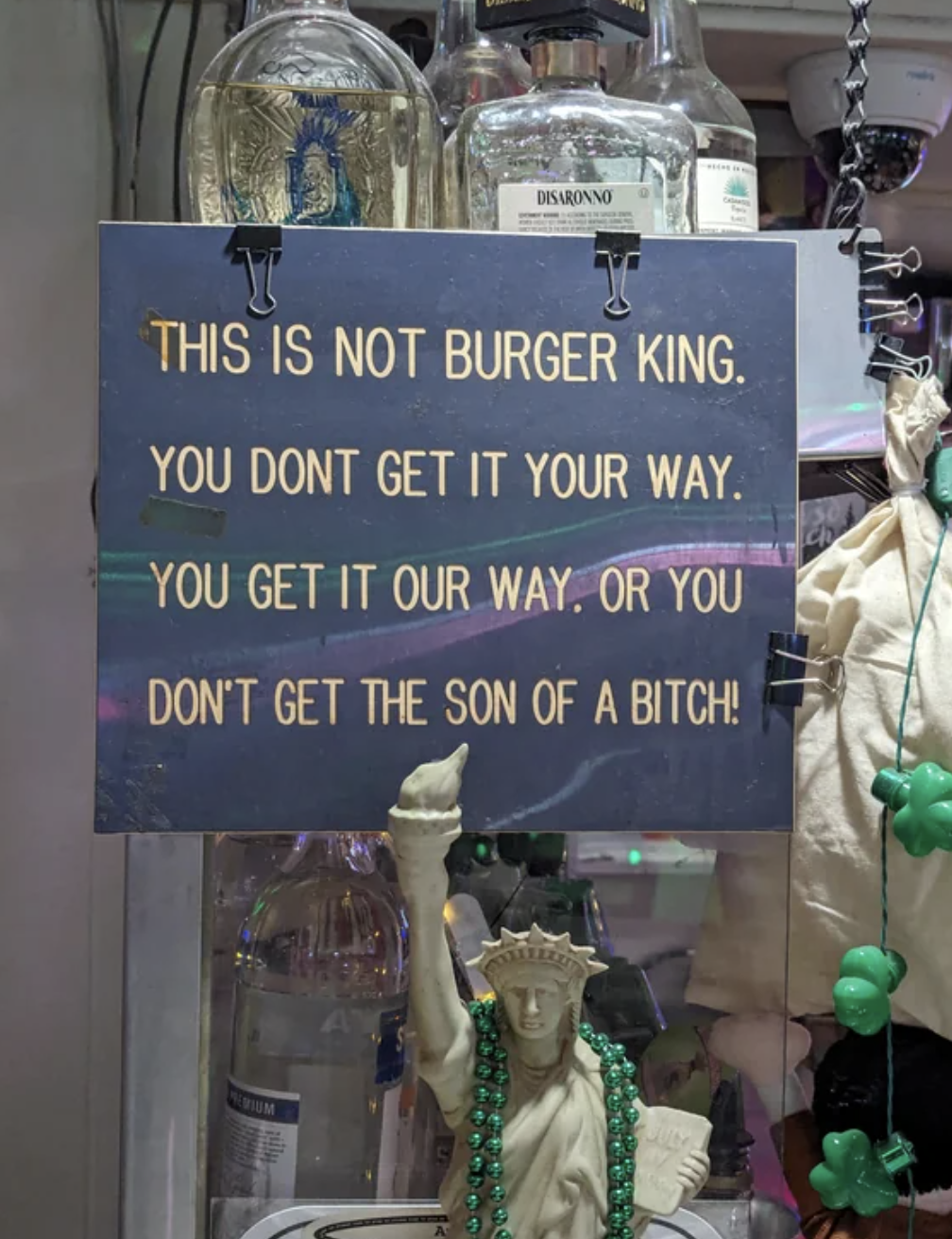 Sign reads &quot;This is not Burger King. You don&#x27;t get it your way. You get it our way or you don&#x27;t get the son of a bitch!&quot; surrounded by bottles and decor