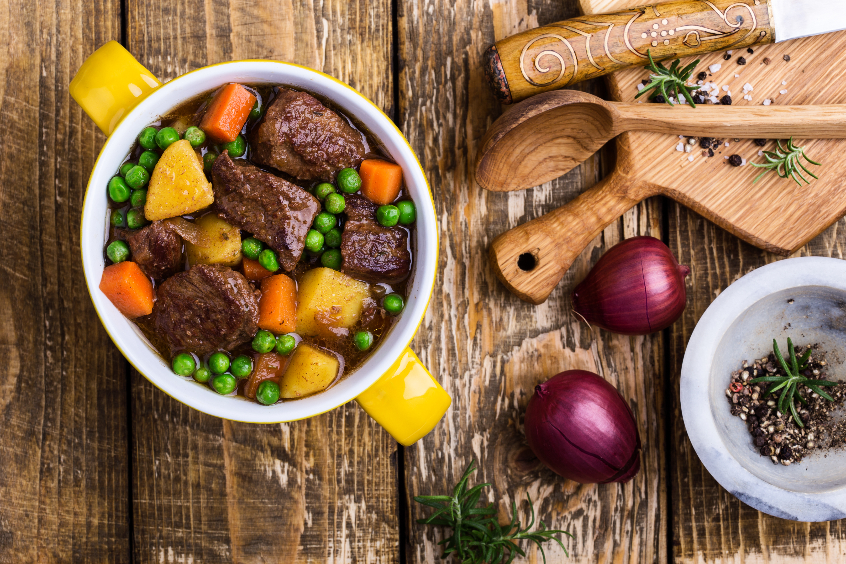Bowl of beef stew with potatoes, carrots, and peas, alongside cooking utensils and herbs