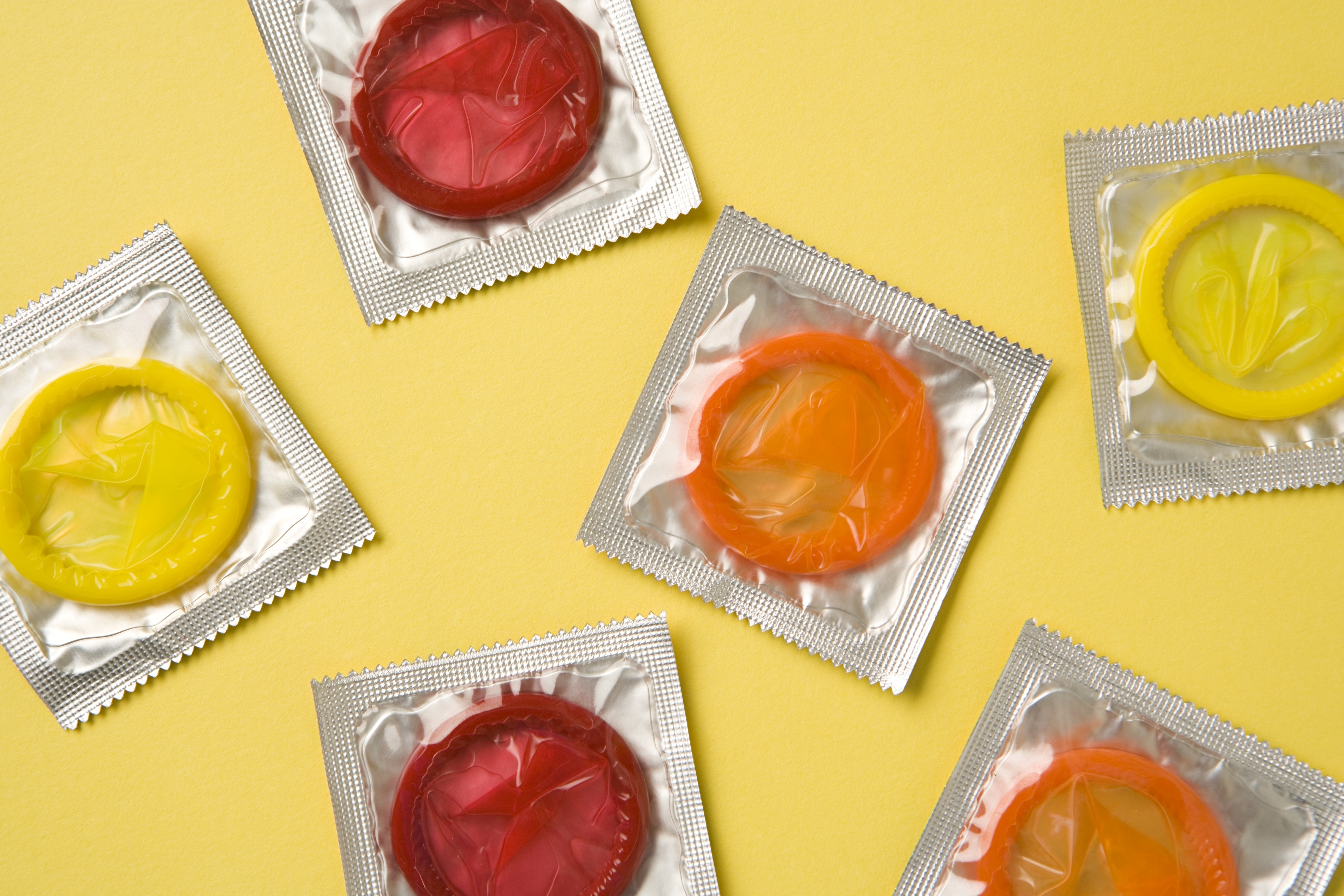 Six colorful condoms in packaging scattered on a yellow background