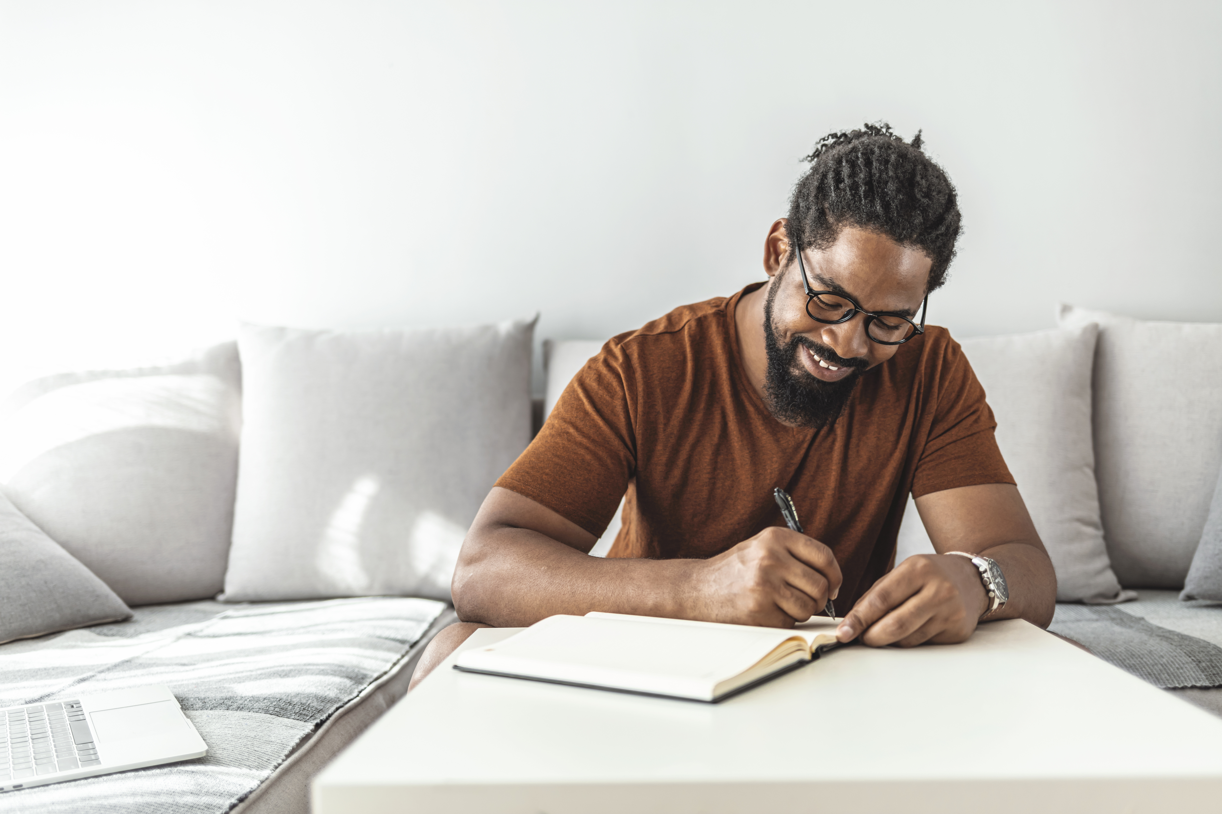 Man writing in notebook on couch with laptop nearby