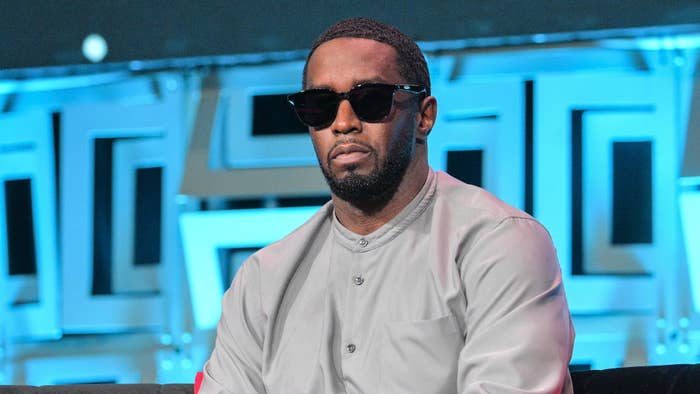 Diddy in a buttoned-up shirt and shades, seated with a conference backdrop