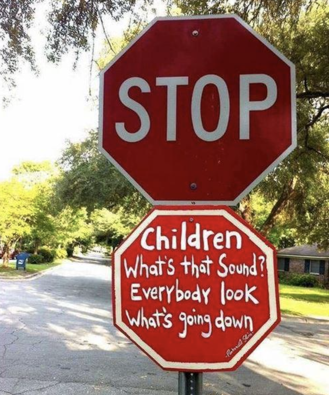 Stop sign with added text &quot;Children What&#x27;s that Sound? Everybody look What&#x27;s going down&quot; on a tree-lined street