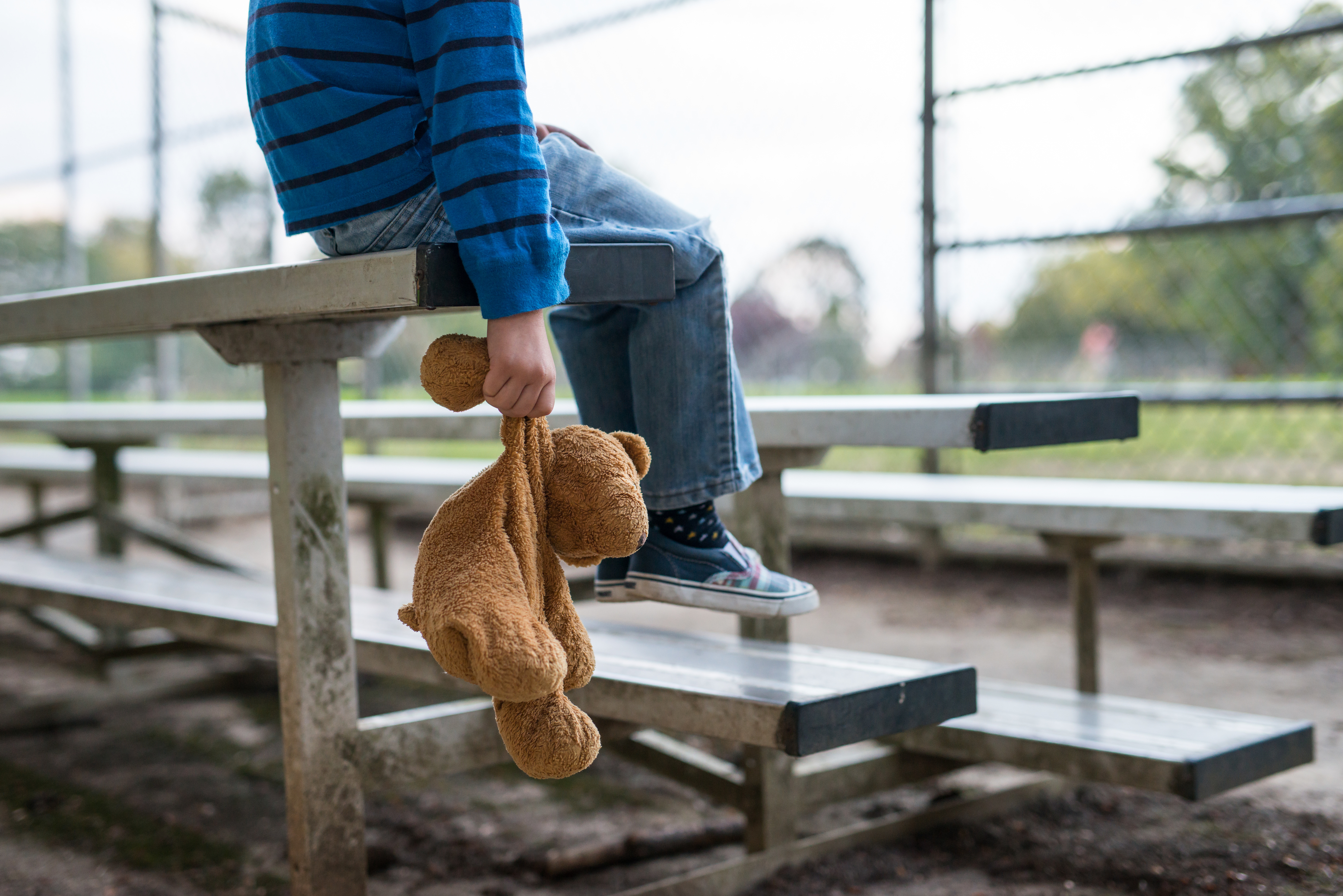 Child sitting on a bench holding a teddy bear, in a park setting, facing away from the camera