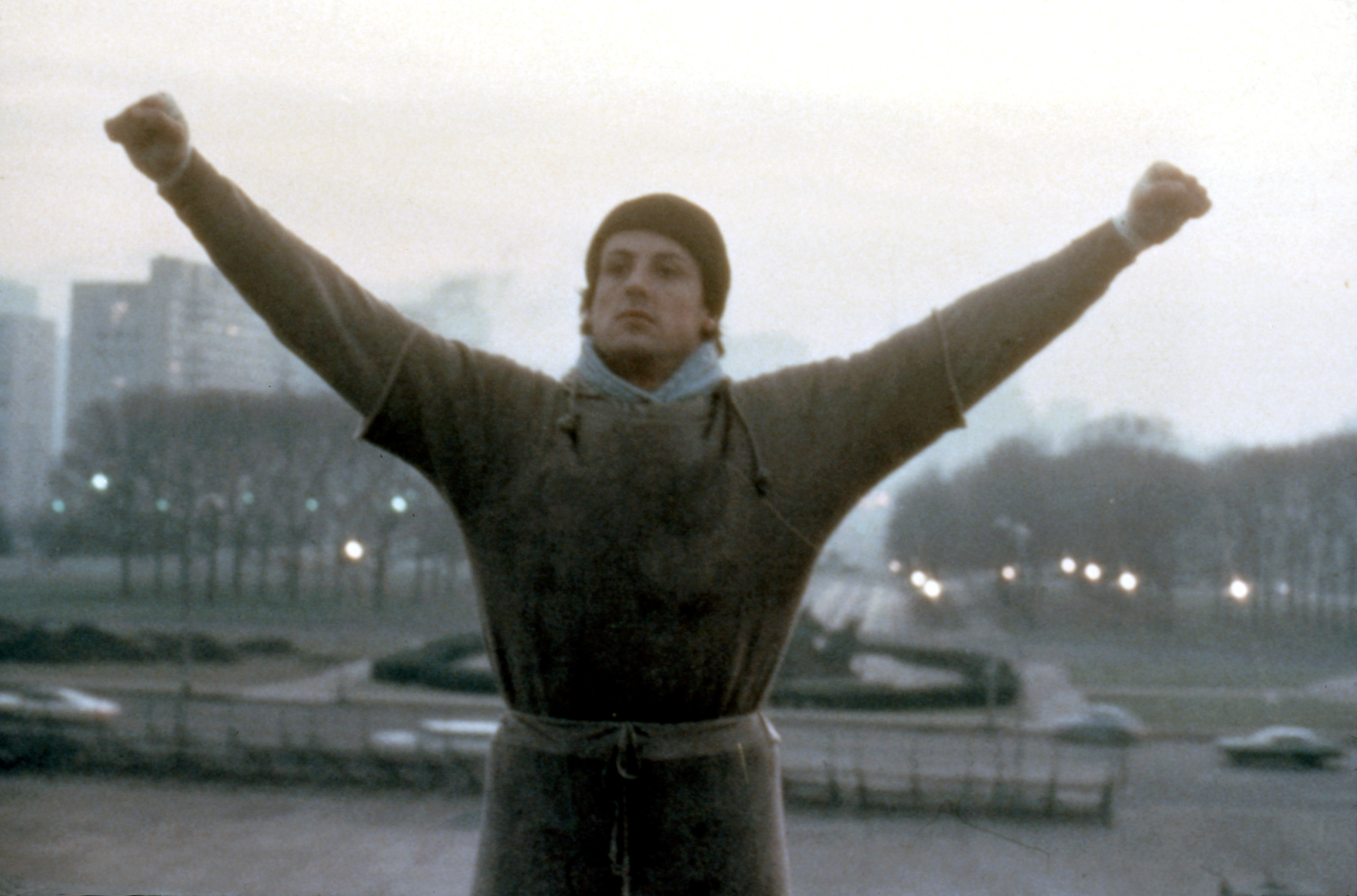 Sylvester Stallone as Rocky Balboa in a gray sweatshirt with arms raised in triumph