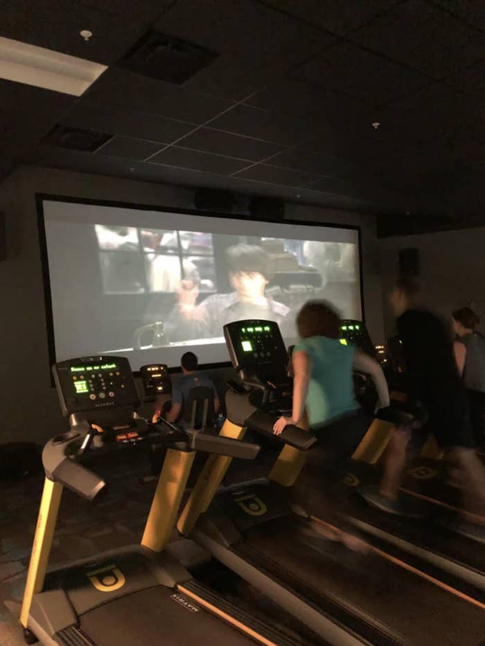 People exercising on treadmills in a dark room while watching a movie on a large screen