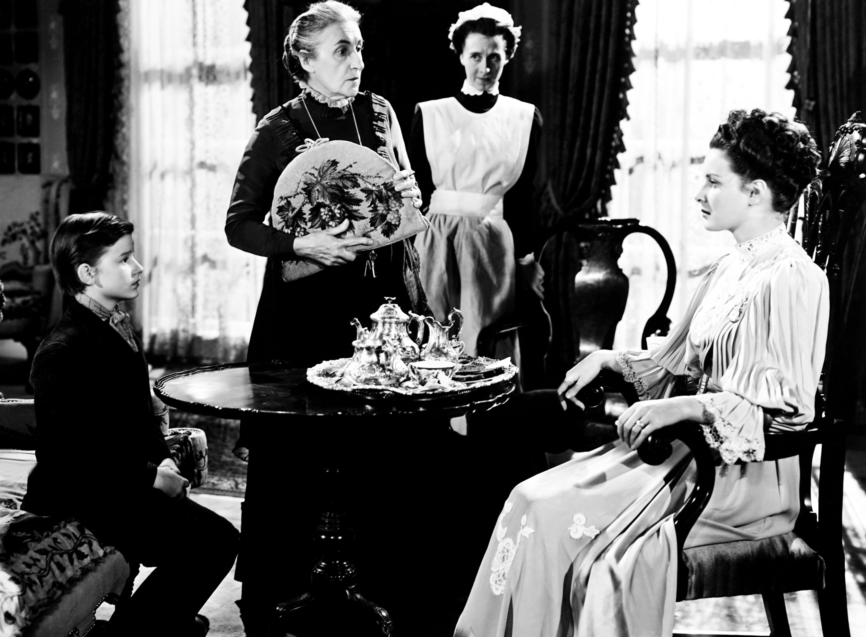 Vintage film scene with four actors in period clothing, indoors, with one seated, a boy listening, and two women standing, one holding a fan and one in a maid uniform