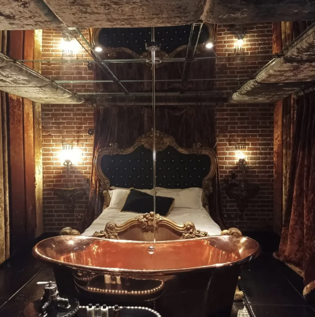 Ornate bedroom with copper bathtub, large bed with canopy, and exposed brick walls
