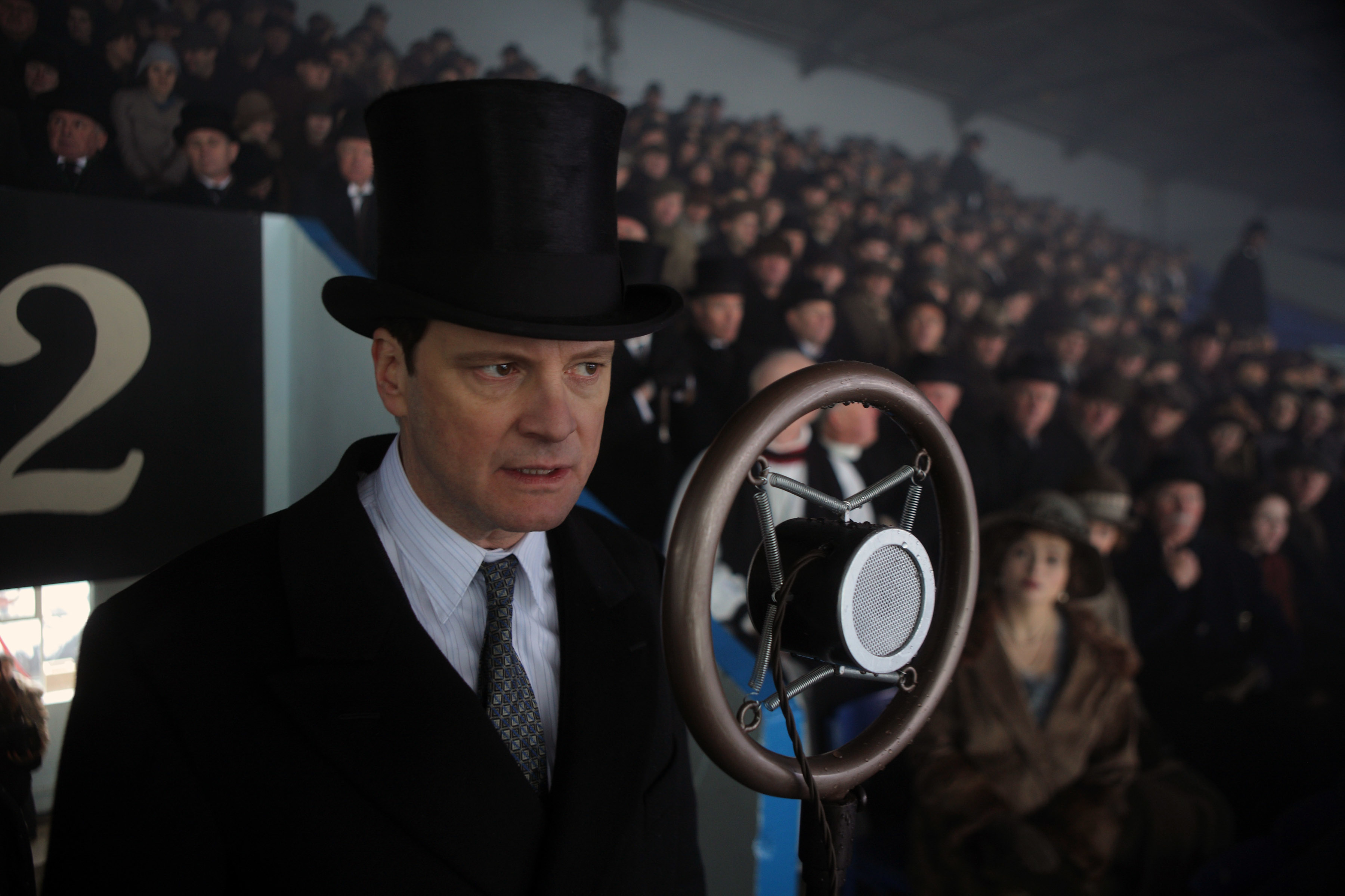 Colin Firth as King George VI, in a suit and tie and top hat, speaks into a vintage microphone