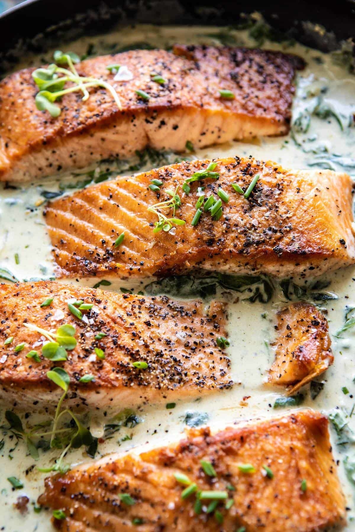 Four cooked salmon fillets in a creamy sauce garnished with herbs in a skillet