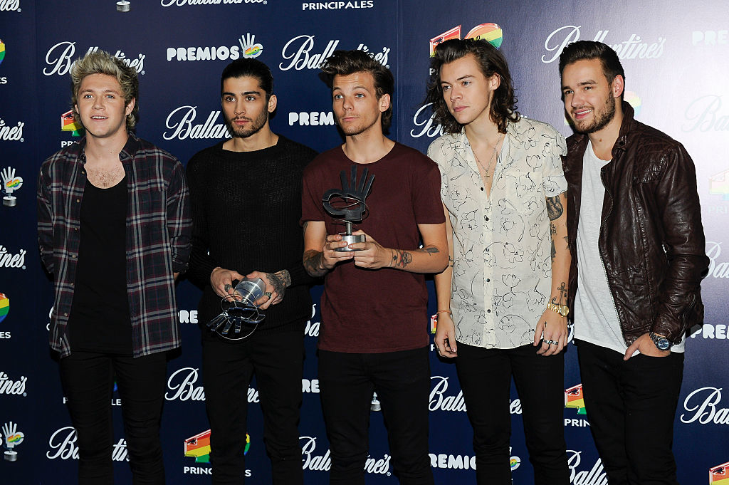 Five members of One Direction posing with awards, in stylish casual wear