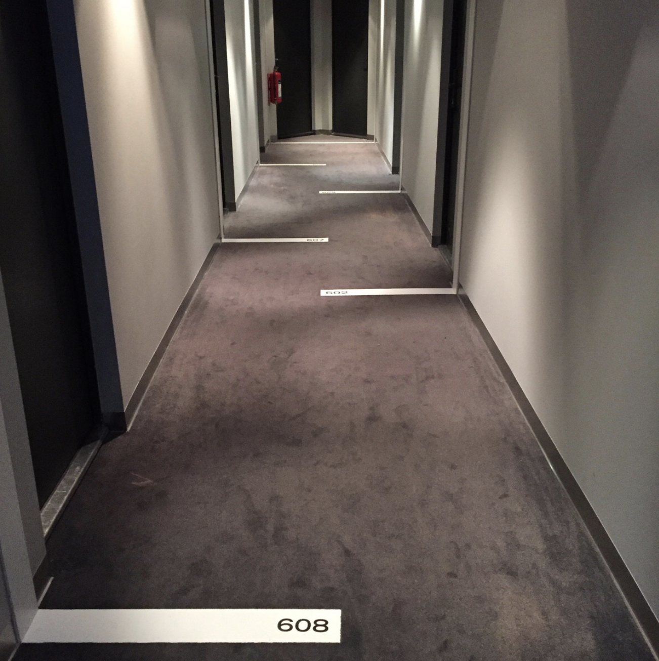Narrow hotel hallway with room numbers visible, leading toward a distant exit sign