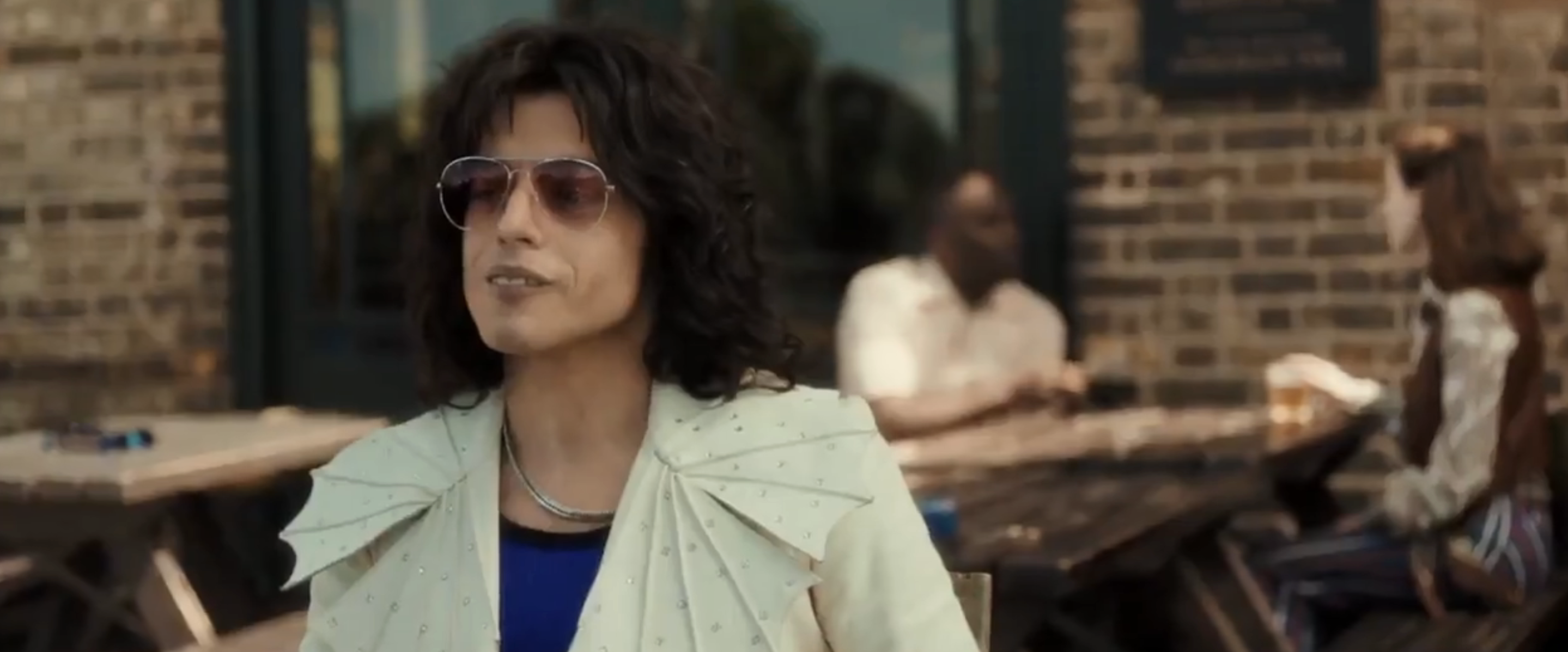 Rami Malek as Freddie Mercury in a patterned blazer and sunglasses seated outside with people in background