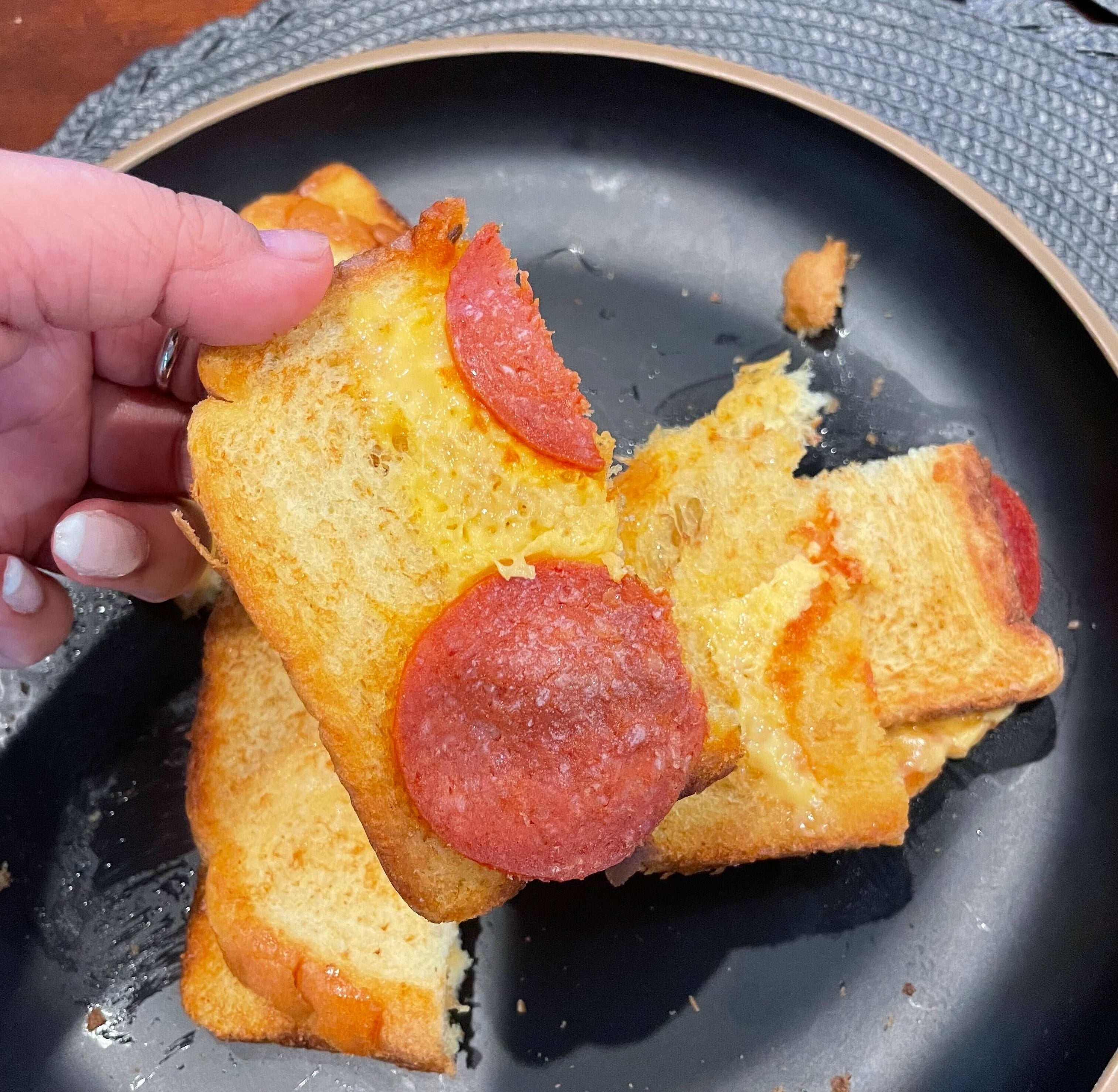 A hand holding a slice of pepperoni pizza toast over a black plate with crumbs