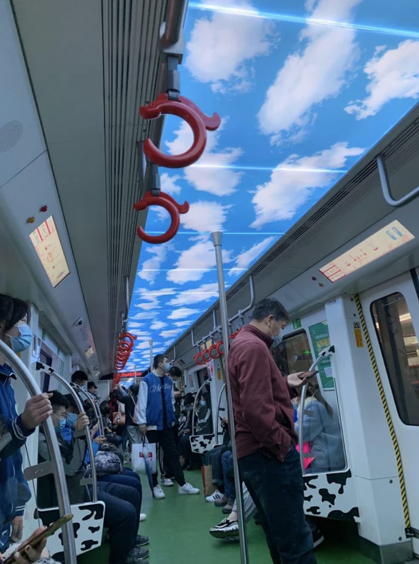 Commuters stand and sit inside a train with overhead handles, one person is reading a newspaper