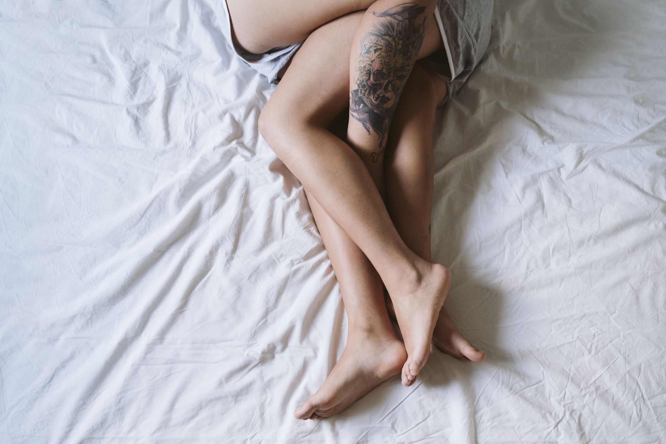 Two people hugging knees, lying on bed, one with floral tattoo on right thigh