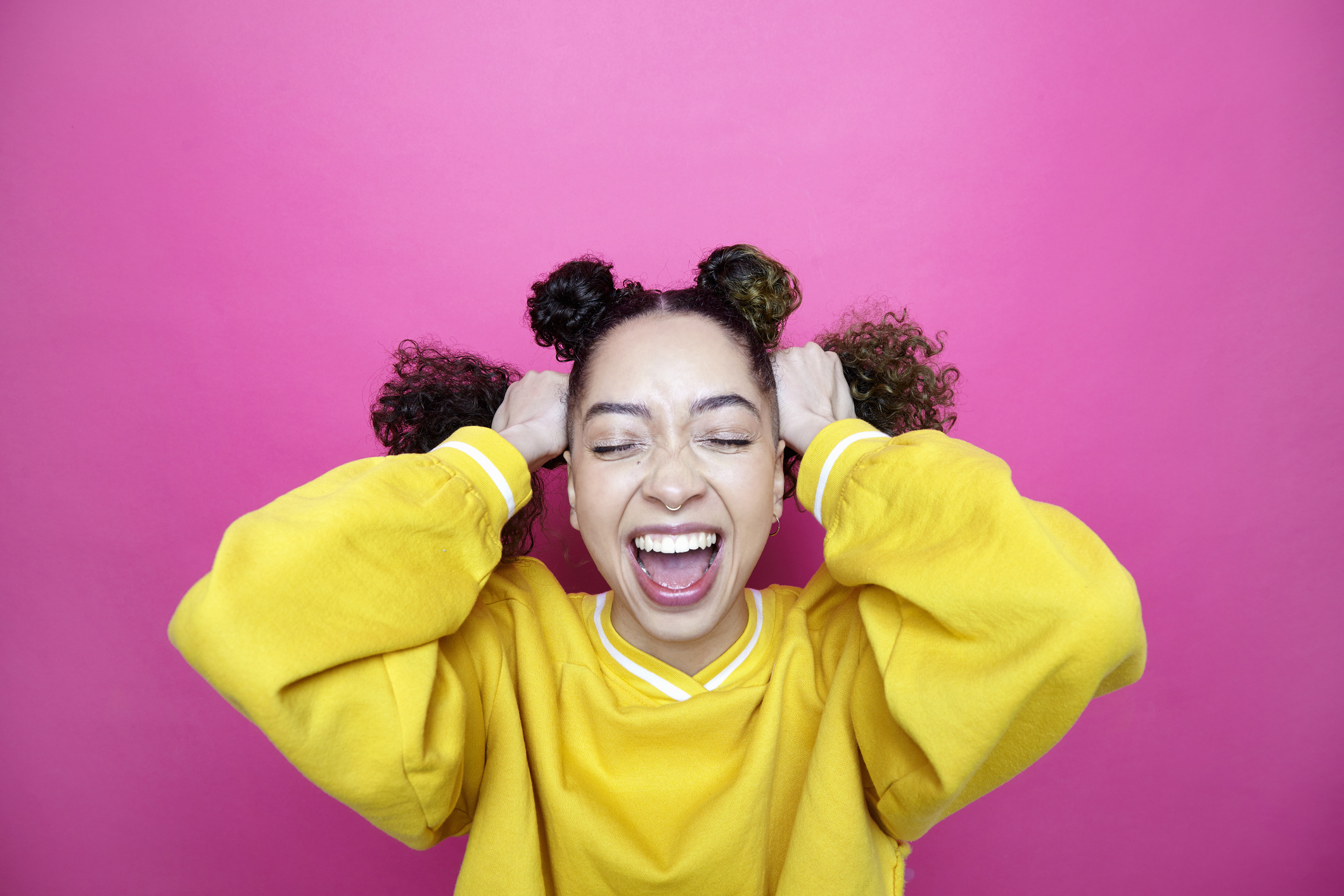 Woman with hair in buns, yellow top, laughing with hands on head against pink background