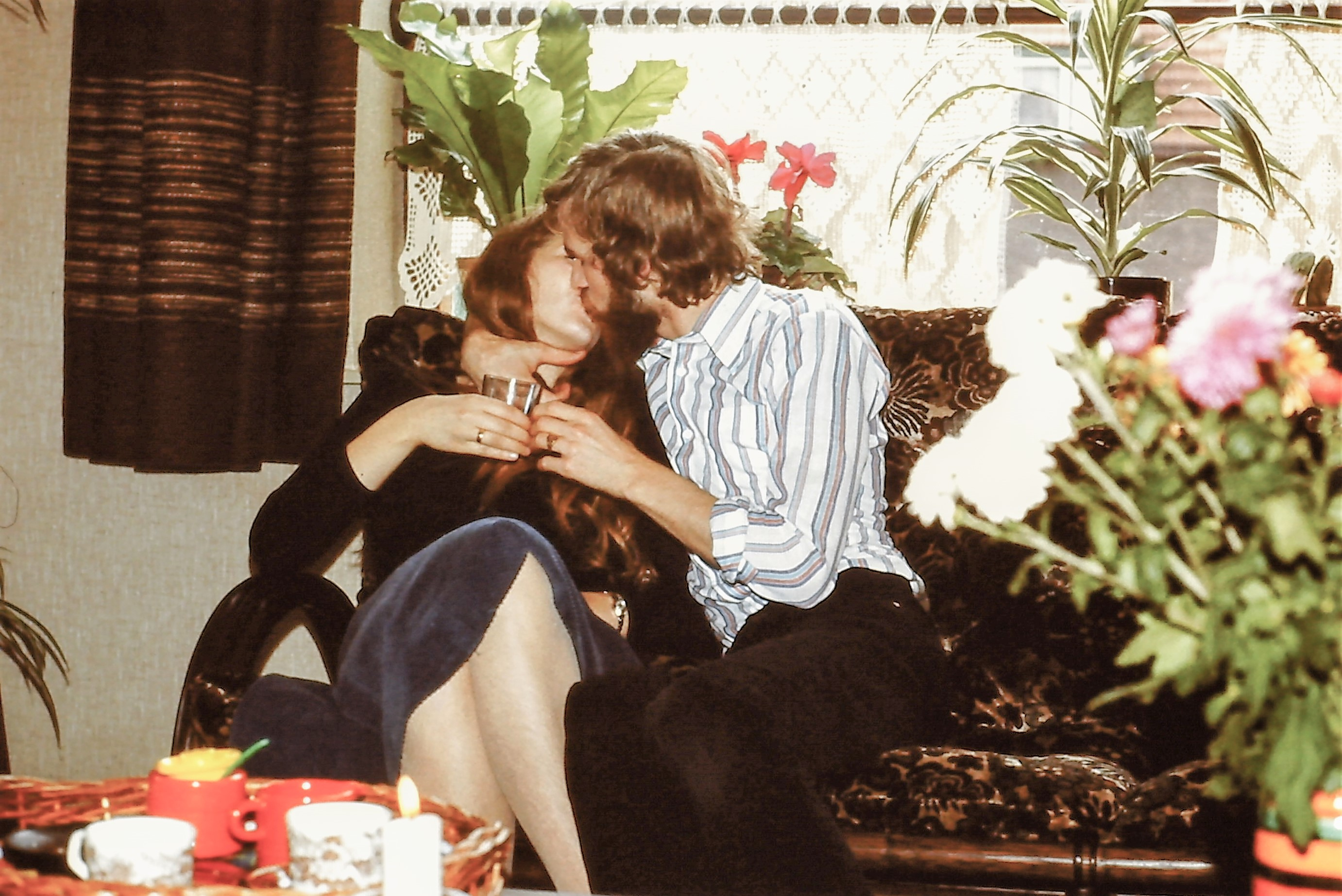Two people sharing a kiss on a couch, surrounded by houseplants and a window in the background