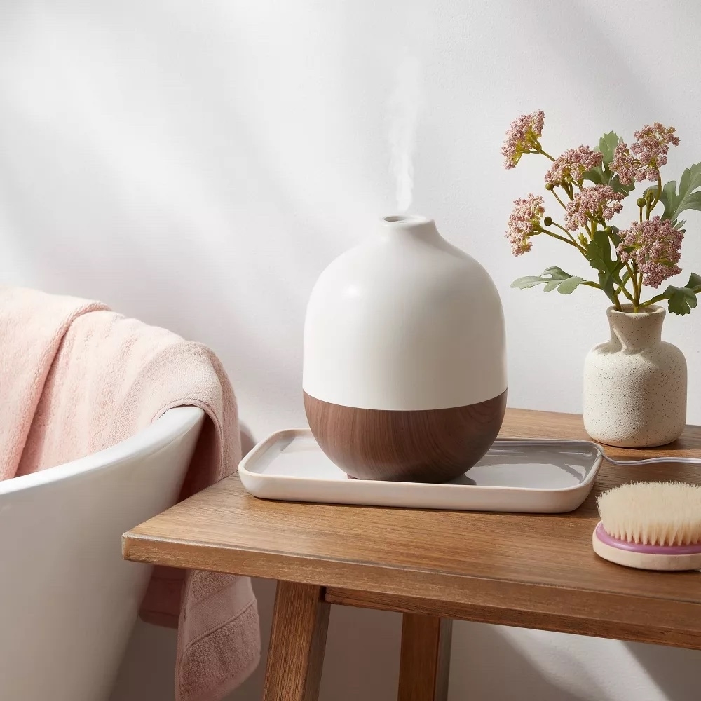 Ultrasonic diffuser on a wooden stool next to a vase with flowers and a brush, emitting steam for a soothing atmosphere