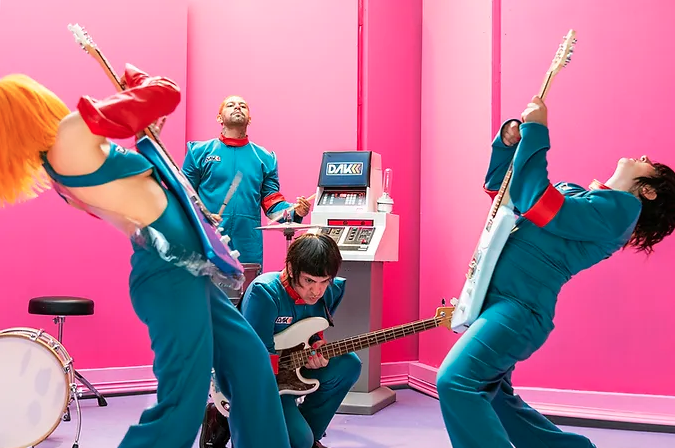 Banda de cuatro miembros en uniformes azules tocando rock con entusiasmo en un cuarto rosa