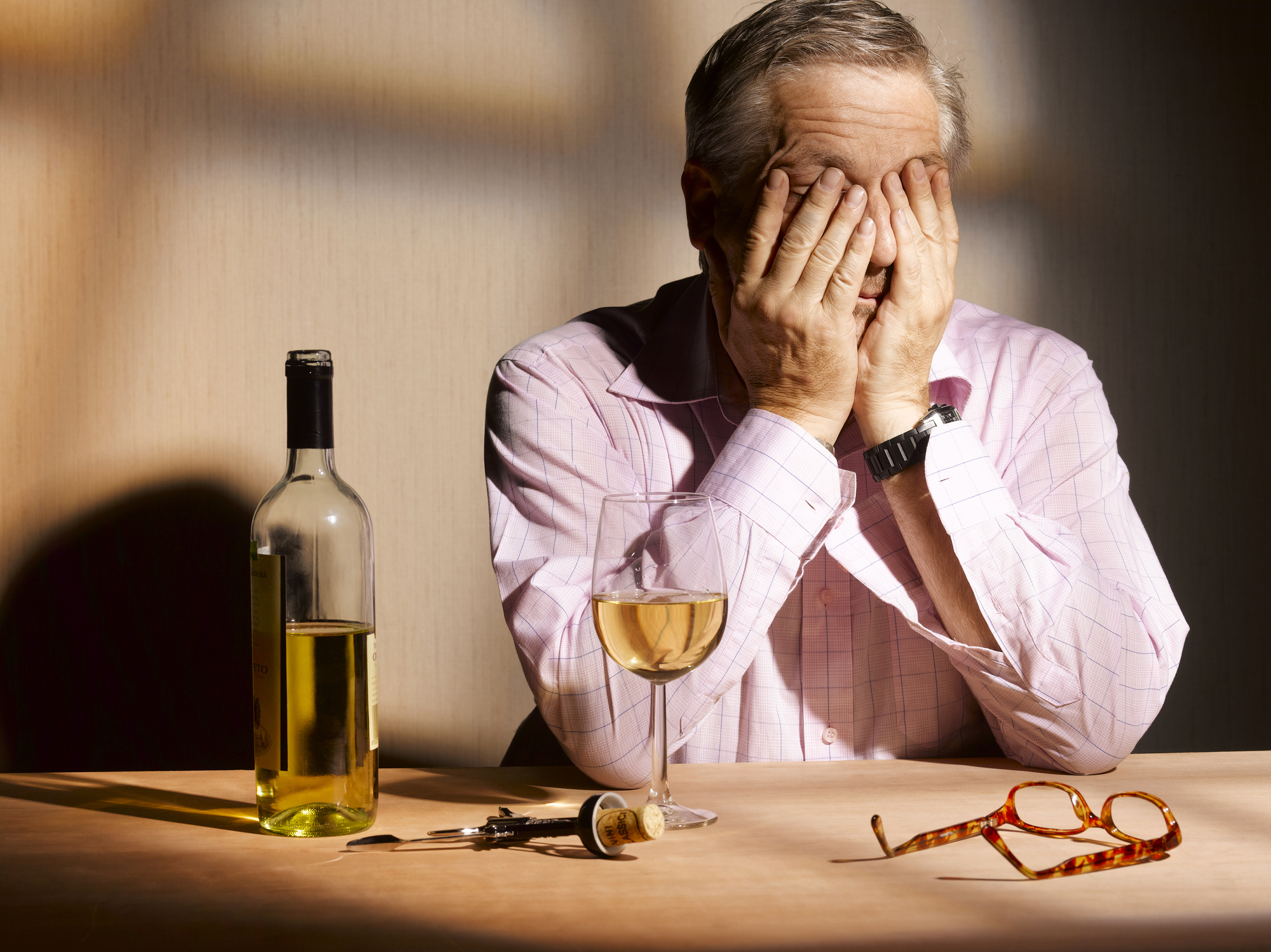 A man covering his face as a glass and bottle of wine sit on the table in front of him