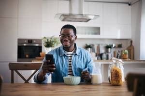 Man sitting at dinner table in the kitchen with a bowl of cereal smiling at his phone
