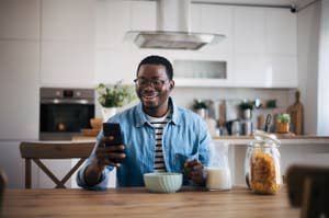 Man sitting at dinner table in the kitchen with a bowl of cereal smiling at his phone