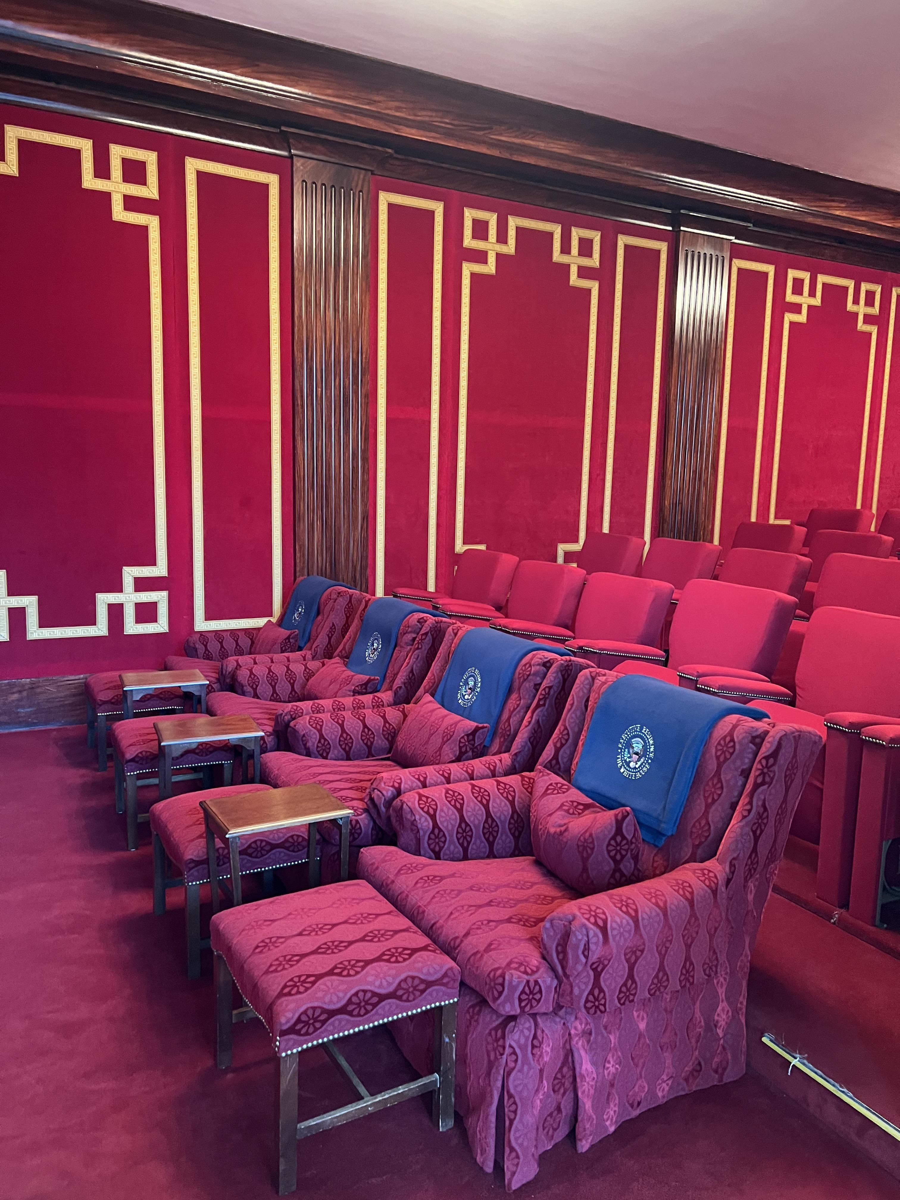 Red rug and rows of seats; the first row has footrests/ottoman tables and presidential seals on the top of the chair