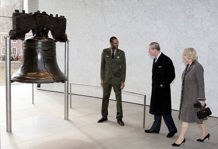 A man in a military uniform with King Charles III and Queen Camilla standing near the bell, which is on a pedestal and stands a bit higher than they are