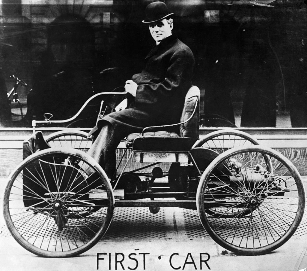 A man with a top hat sitting in a car that looks more like a four-wheel bicycle/carriage