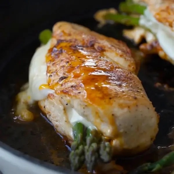 Closeup of cooked chicken stuffed with cheese and asparagus, topped with sauce, in a pan
